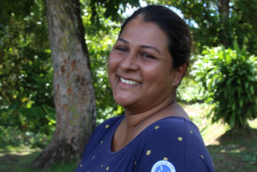 A Fijian woman standing side on smiles with trees in the background