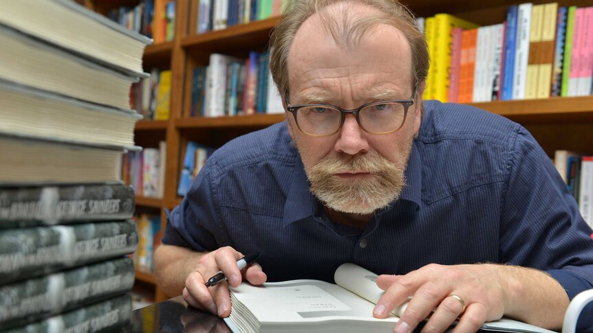 Colour photograph of author George Saunders signing a stack of books in a bookstore.