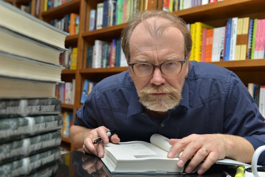 Colour photograph of author George Saunders signing a stack of books in a bookstore.
