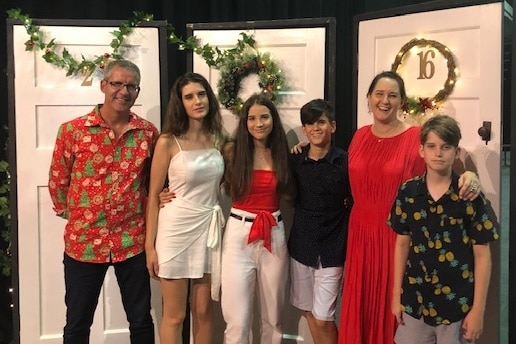 Six people stand in front of three white doors with Christmas decorations, smiling.