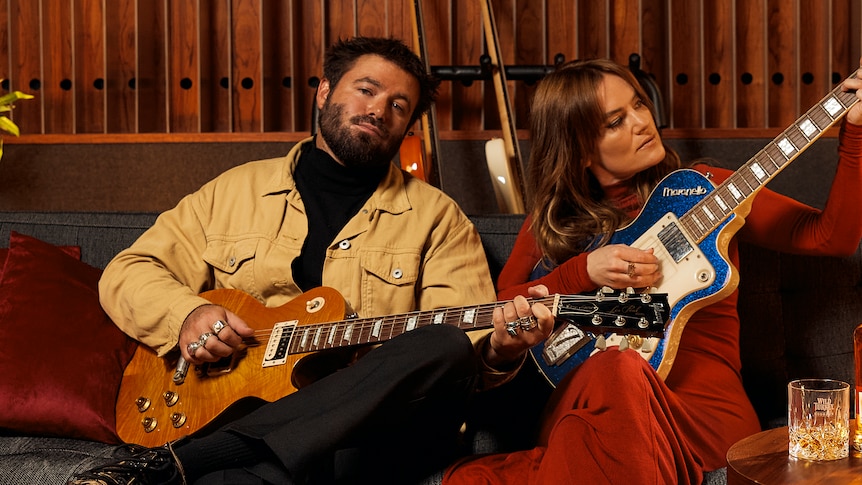 A man and a woman sit on a couch looking at the camera. They both hold guitars.