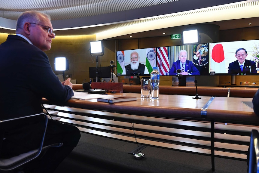 A middle-aged man in a suit at table talks to three elderly men via monitors in wooden boardroom.