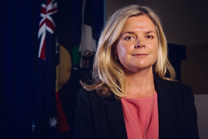 Dr Katrina Sanders in a dark room. She has an Australian flag and an Aboriginal flag behind her.