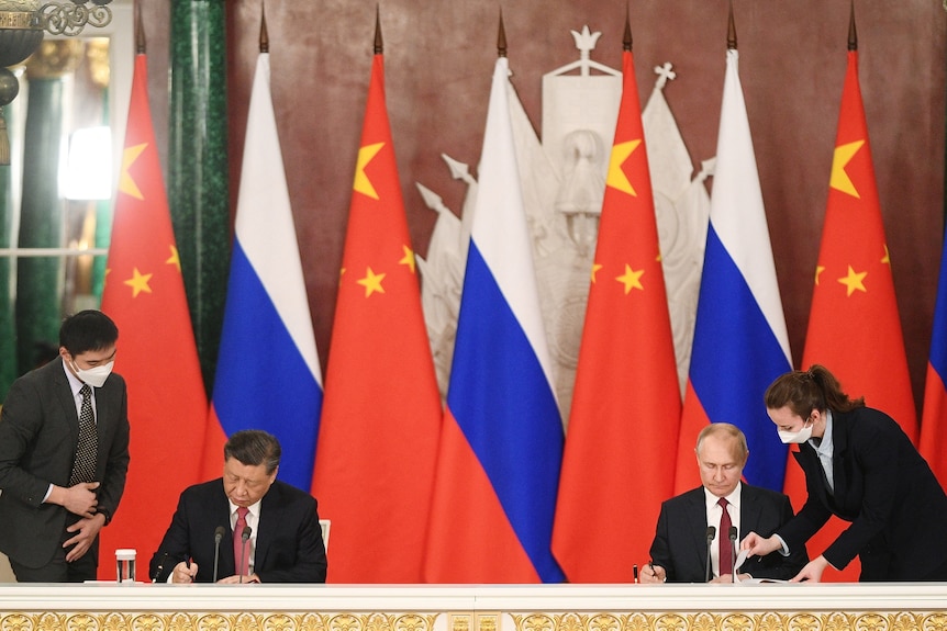 Xi, seated left and Putin sit a distance apart as they sign a document. An aide leans over the shoulder of each