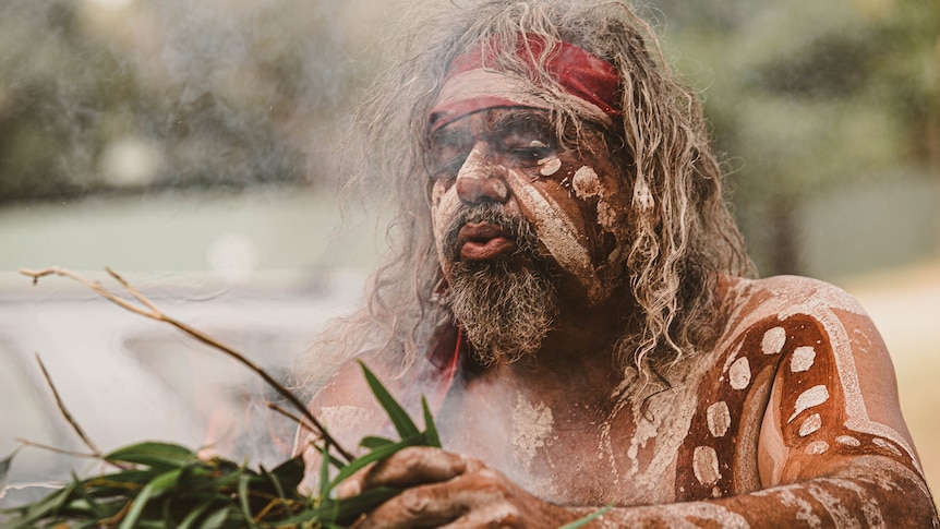 An Aboriginal main painted in ochre, holding smoking gum leaves.