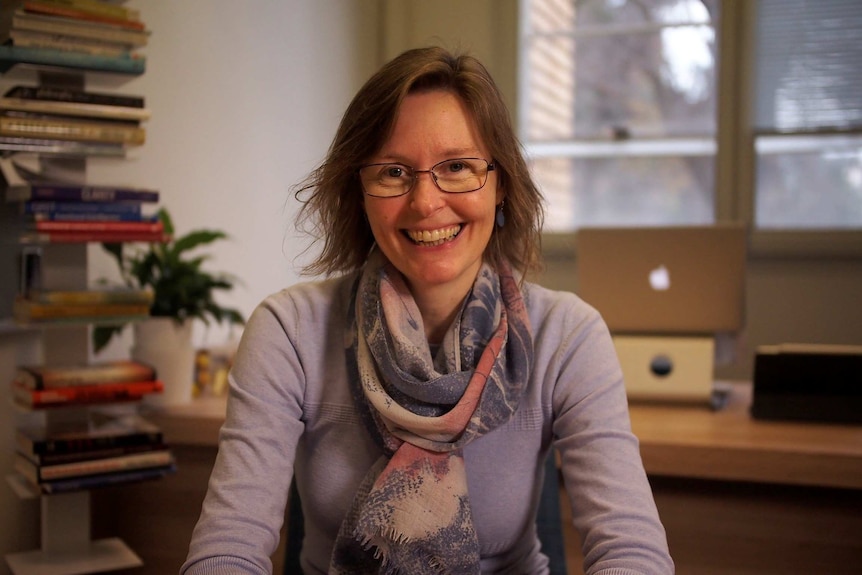 In a dimly-lit office surrounded by books, you view a woman with glasses smiling at the camera.