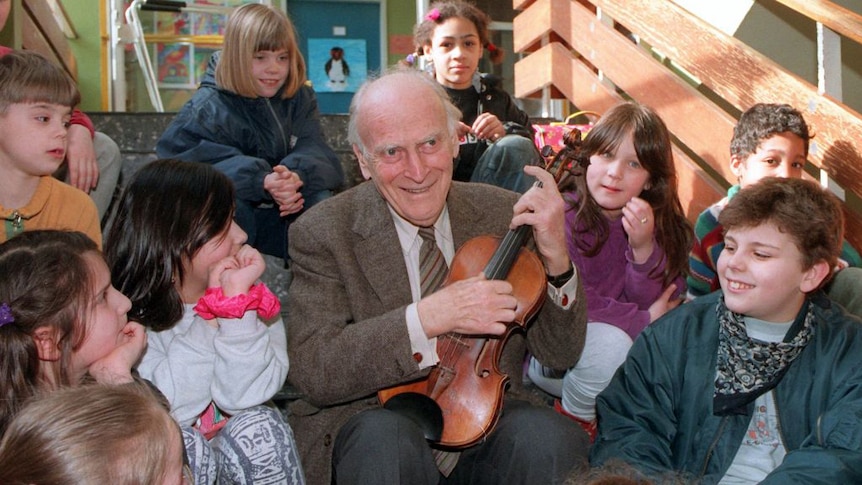 Yehudi Menuhin seated on some stairs holding a violin and surrounded by young children.
