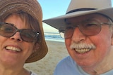 A middle-aged woman and older man, both in hats and glasses, smile at camera on beach with blue sky.