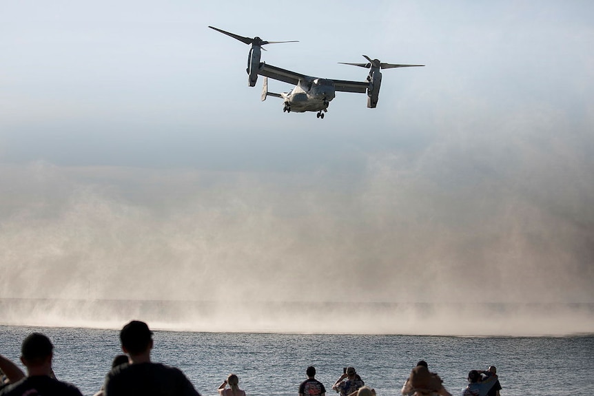 A large military aircraft hovering above water near the beach, with spectators below.