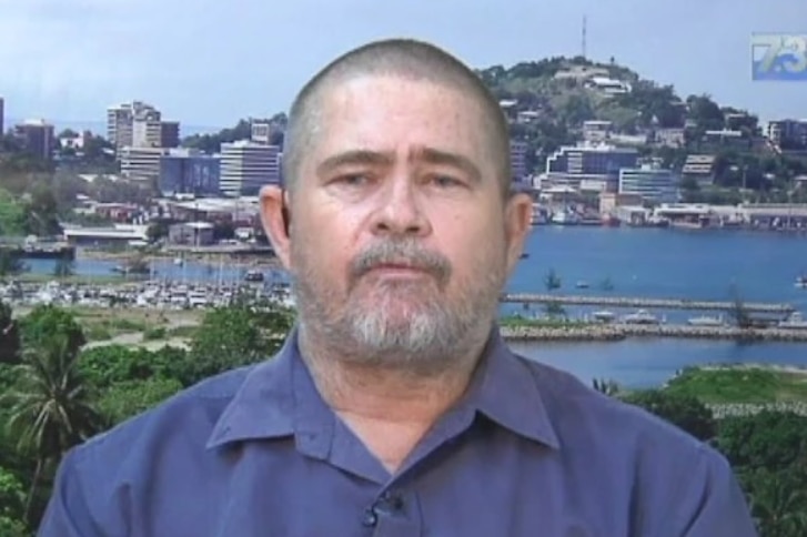 A man with a slight grey beard and short hair in front of a screen displaying the city of PNG