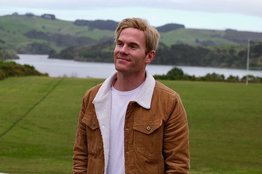 A man with blonde hair and a corduroy jacket with wool lining stands on a rugby field.