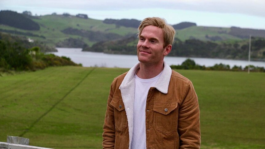 A man with blonde hair and a corduroy jacket with wool lining stands on a rugby field.