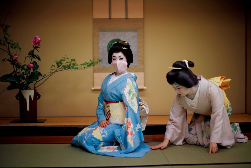 A geisha in a blue kimono and face mask kneels on a mat while another woman smooths out her dress