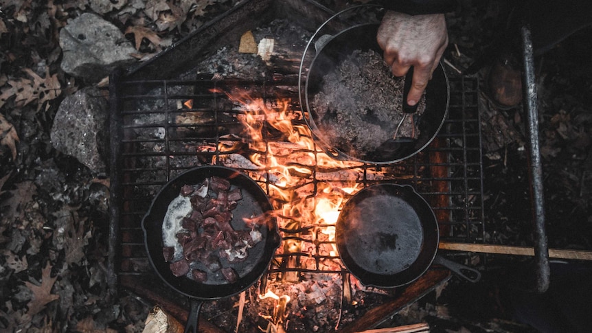Person cooking in cast iron pots over coal fire on ground.