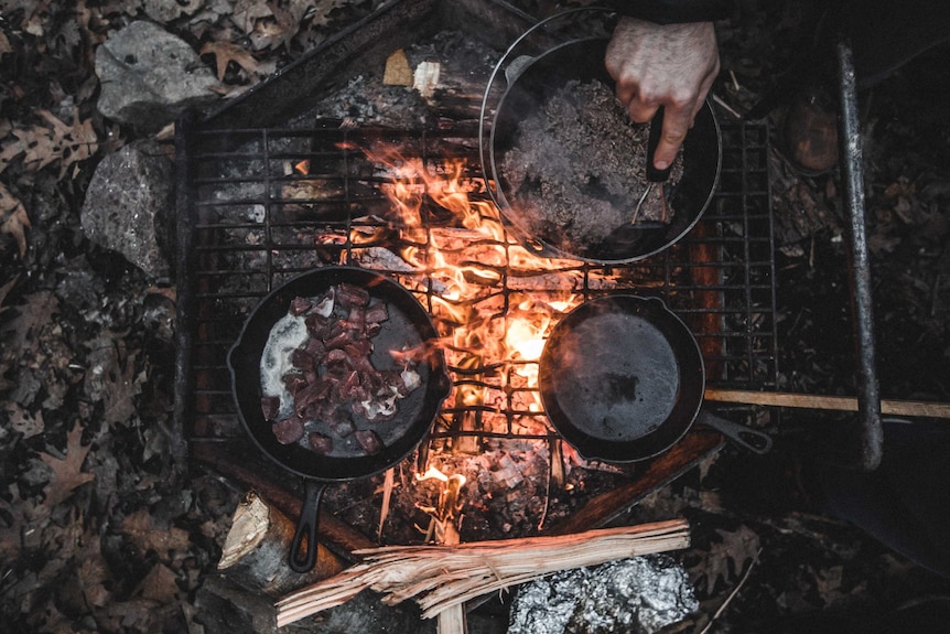 Person cooking in cast iron pots over coal fire on ground.