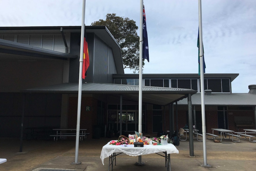 Three flag poles with flags at half mast and a table containing floral tributes and bunches of flowers.