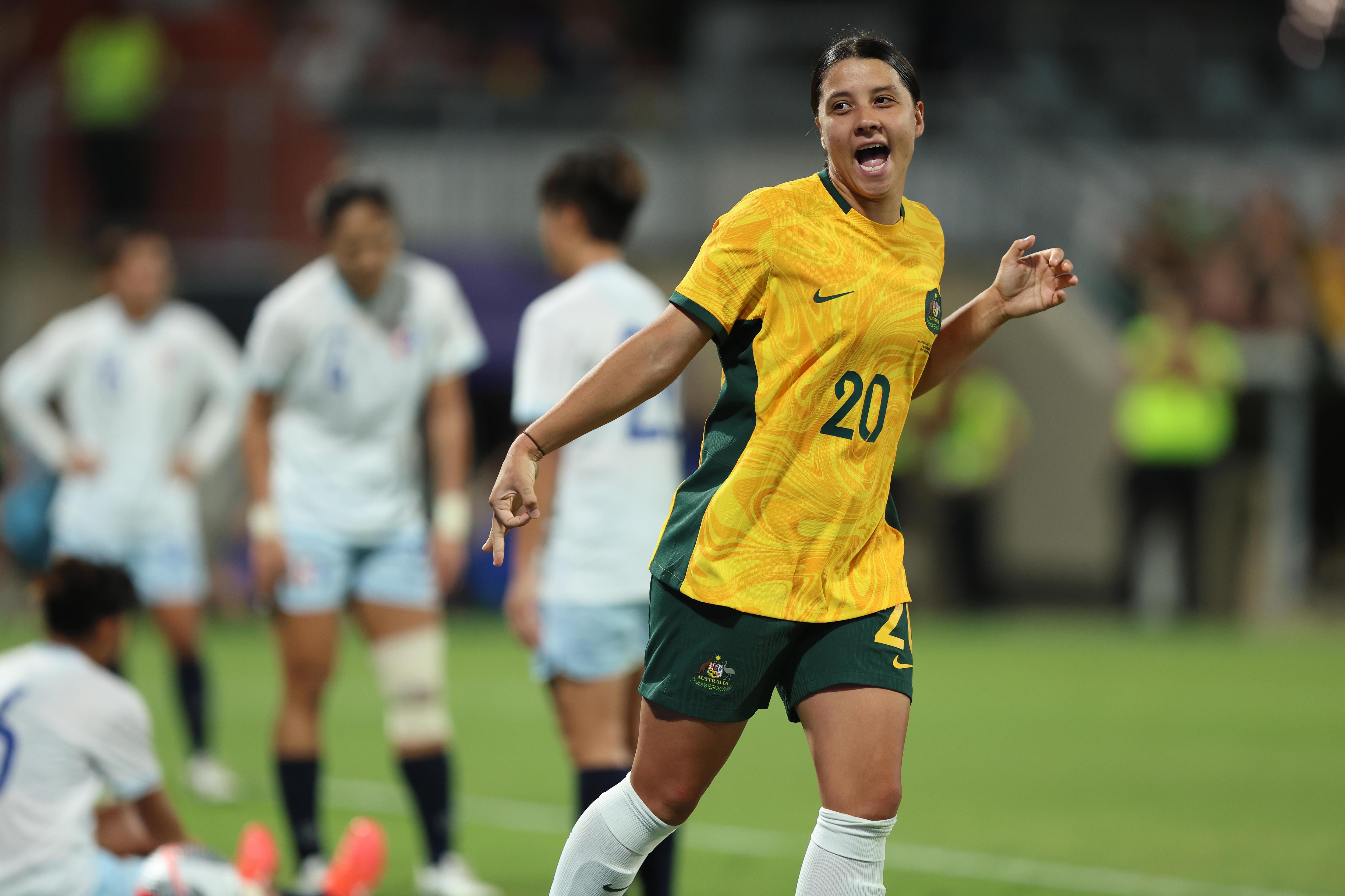 A female footballer wearing gold, running away in celebration, smiling, after scoring