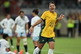 A female footballer wearing gold, running away in celebration, smiling, after scoring