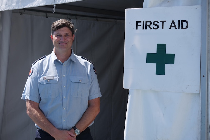 A man in a blue paramedics t-shirt standing in a tent, next to a sign that says "FIRST AID"