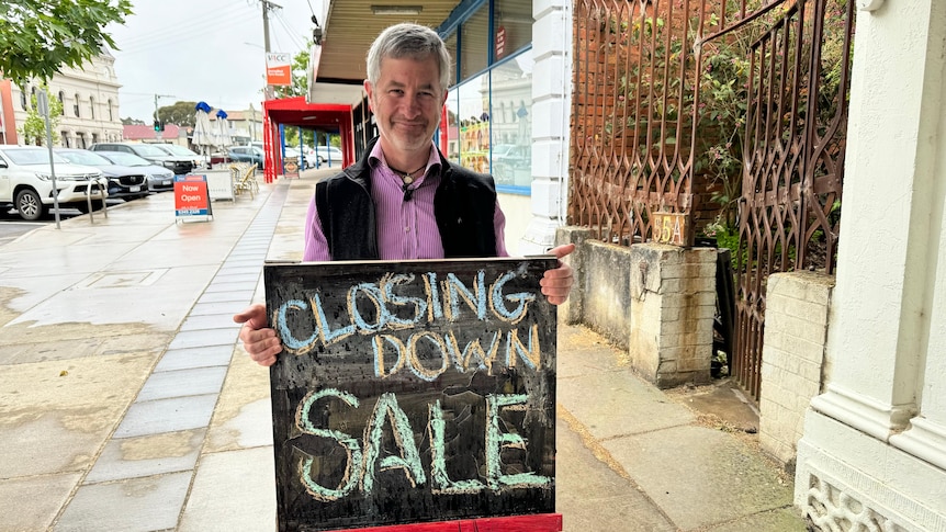 A man standing ona footpath outside a shop holding a sign that says 'closing down sale'.