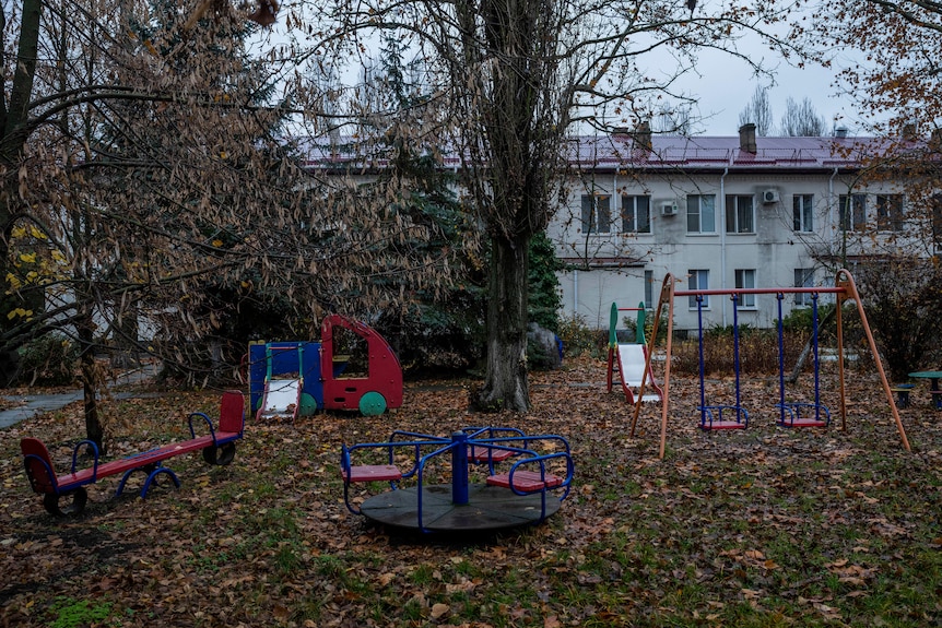 Children's play equipment, including a swingset, outside on grass covered in leaves with no children playing. 