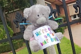 A grey teddy bear is tied to a fence with a colourful sign on it that says 'we're going on a bear hunt'.