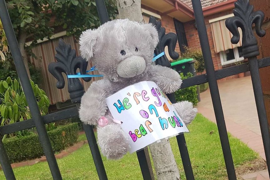 A grey teddy bear is tied to a fence with a colourful sign on it that says 'we're going on a bear hunt'.