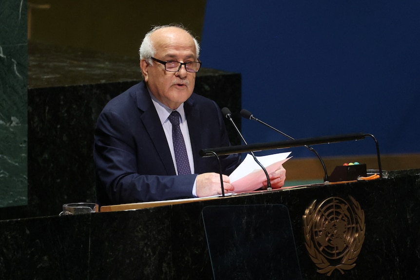 An old man with white hair wearing a dark suit and glasses sitting at a podium with the United Nations logo