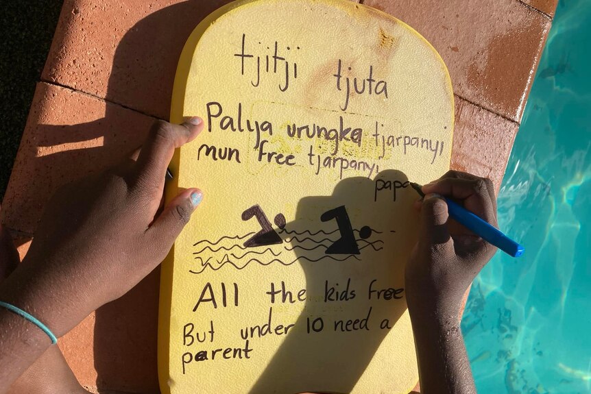 A child writes some of the rules of the pool on a yellow kickboard