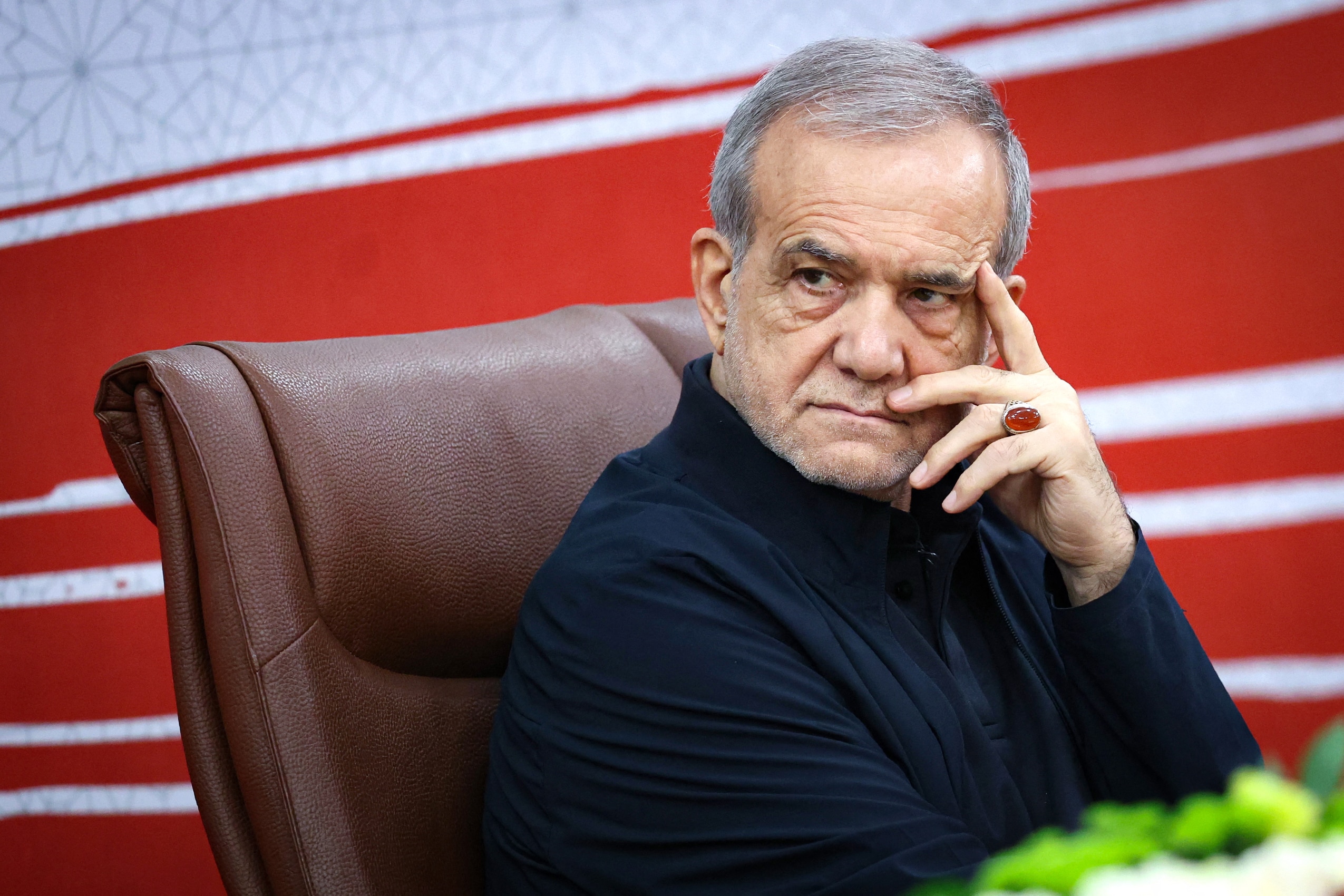 Masoud Pezeshkian sits in a leather chair in front of a red and white background, his hand on his face.