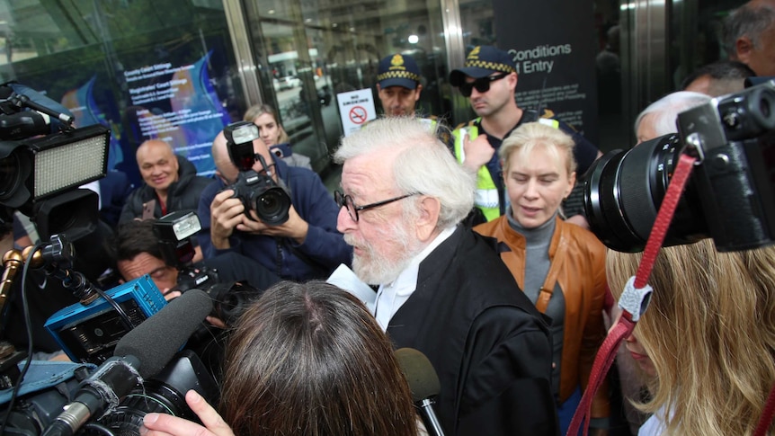 A white-haired man with court robes makes his way through a crowd of media while police look on.