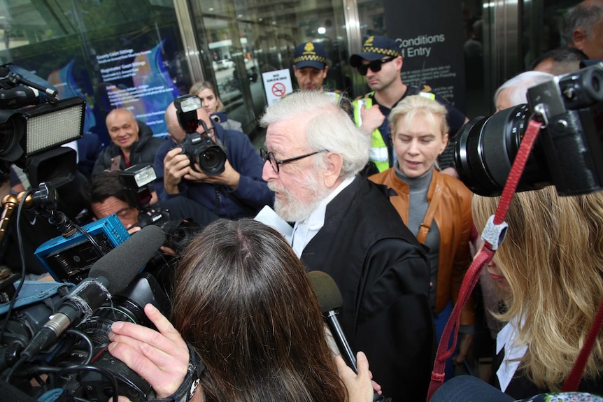 A white-haired man with court robes makes his way through a crowd of media while police look on.