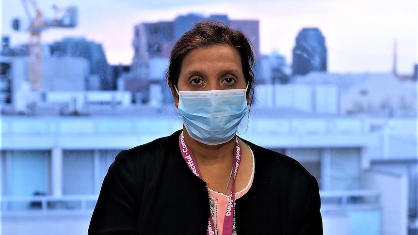 Joy stands wearing a surgical mask, looking to the camera, with a Melbourne city view behind her.