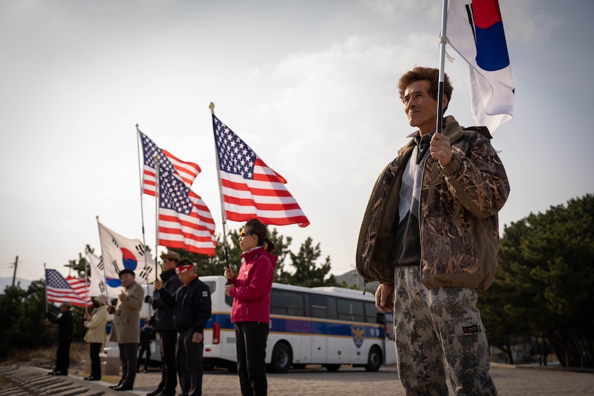 People stand in a line holding up South Korean and US flags, a bus on the road behind