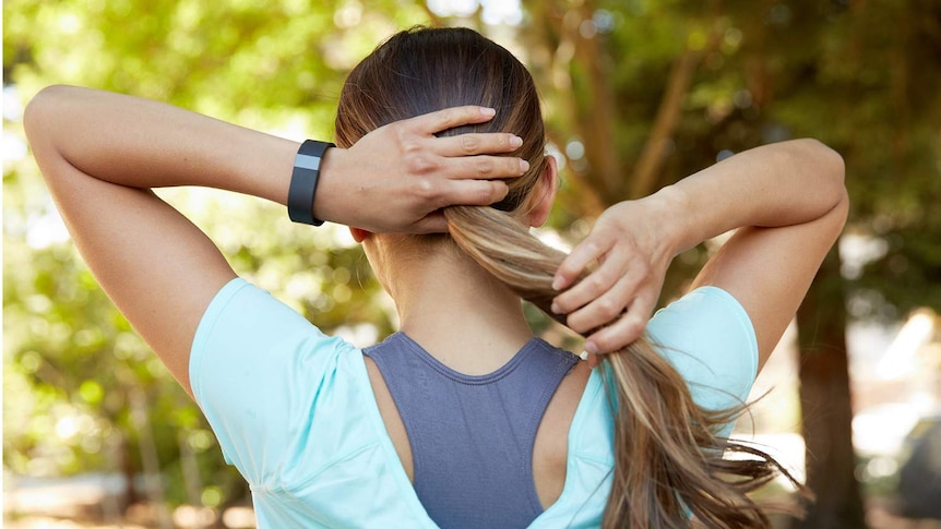 A woman wearing exercise clothes and a fitness tracker pulls her hair back into a pony tail.