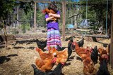Jasmin hugs a chicken on a farm as other chickens at her feet look on.