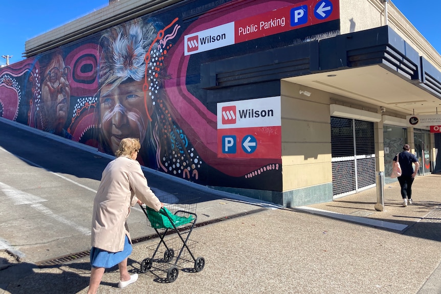 A woman pushes a stroller along a footpath in a sunny day, beside an Aboriginal-design mural.