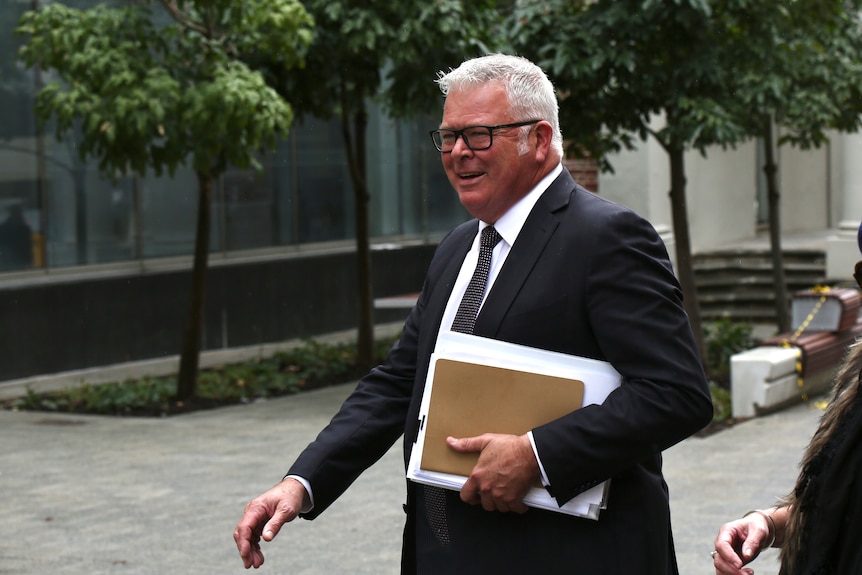 Troy Buswell wearing a black suit and white shirt, holding documents, walking on a footpath outside court.