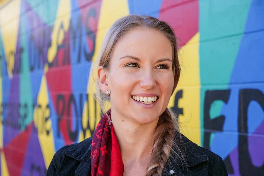 A smiling young woman with long blonde hair in a braid smiles to the left of the camera. She stands in front of a colourful wall