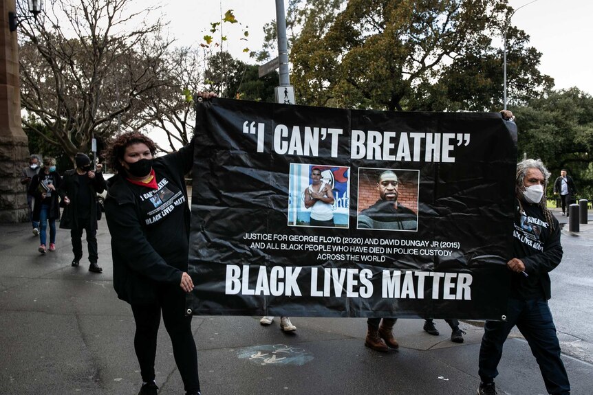 A man and woman walking with a banner held between them which reads I can't breathe, black lives matter.