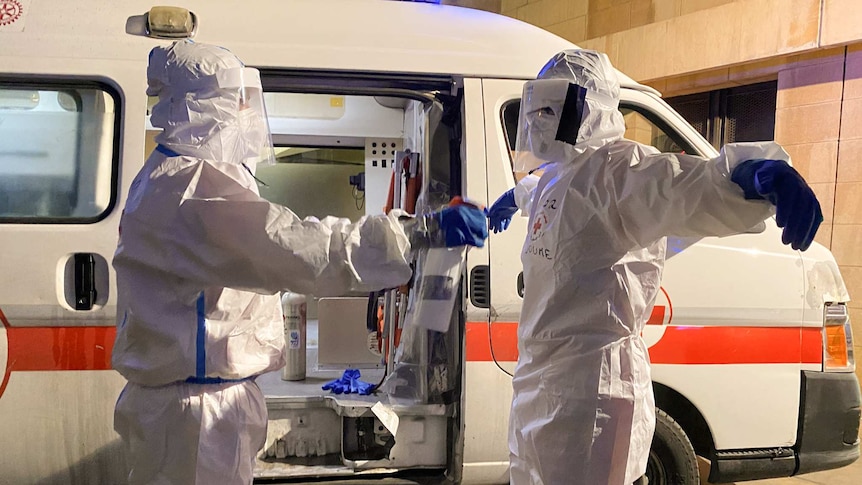 A man in a white PPE suit wearing a mask holds his hands out as he is sprayed by another person outside an ambulance.