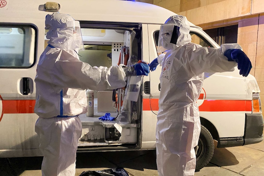 A man in a white PPE suit wearing a mask holds his hands out as he is sprayed by another person outside an ambulance.