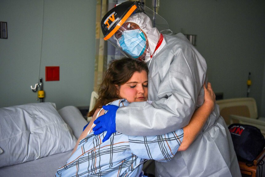 A woman on a hospital bed embraces a health worker in full PPE
