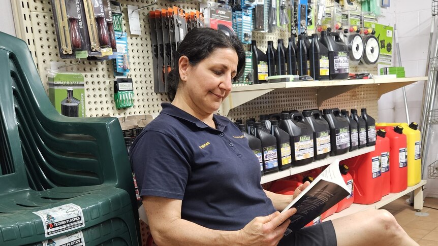 A dark-haired woman sits i9n a hardware store, reading a book.