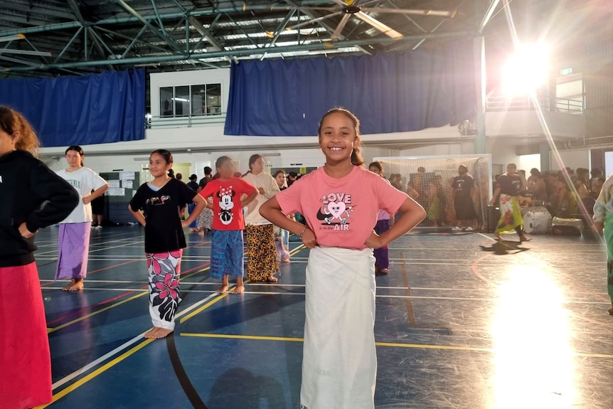 A young Samoan girl wearing pink tee and white ie lava lava stands ready to practise her siva in school hall. 