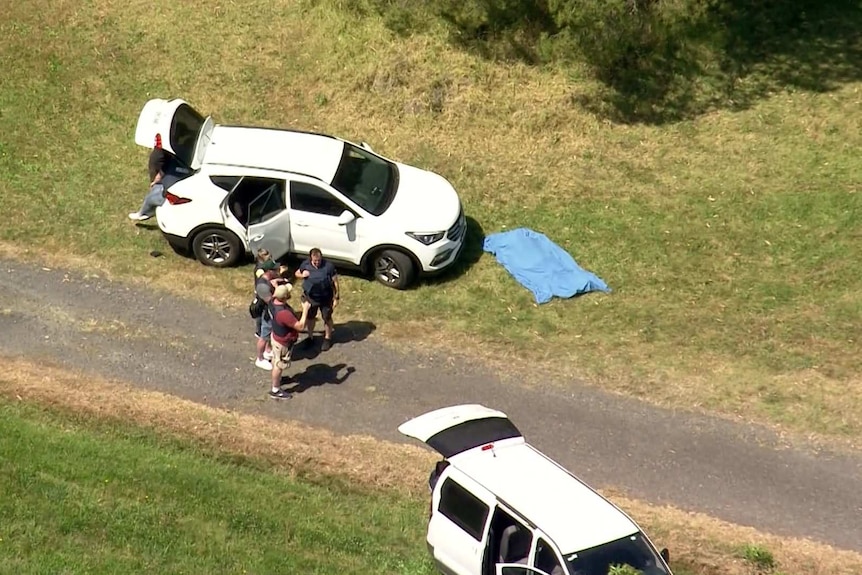 Three men and a woman wearing bullet proof vests stand near a car. A blue blanket covers a body.