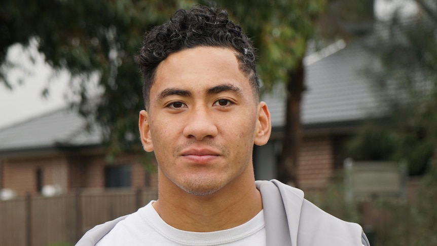 A young man in a Melbourne Storm t-shirt hugs a rugby ball in a carpark.