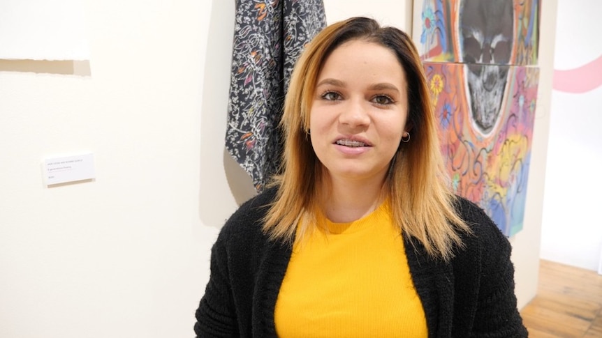 A young Indigenous woman smiles at the camera with artworks hanging behind her