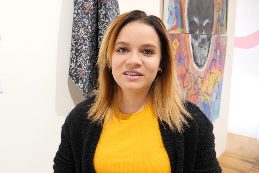 A young Indigenous woman smiles at the camera with artworks hanging behind her