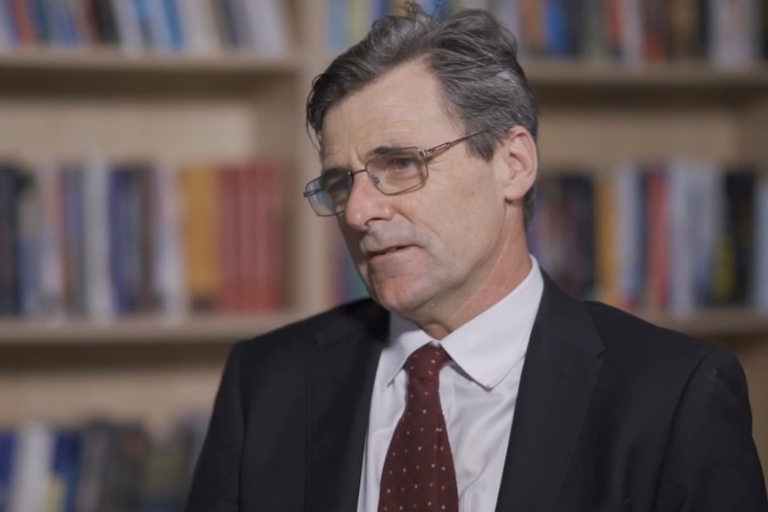 A middle-aged man with grey hair wearing glasses and a suit in an interview with blurred book shelf in background